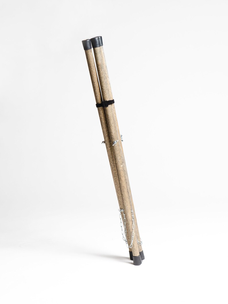 Puede incluir: Un conjunto de tres patas de tr&iacute;pode de madera con puntas de goma negras y una cadena plateada que las conecta. Las patas est&aacute;n plegadas juntas y est&aacute;n listas para ser utilizadas como un soporte.