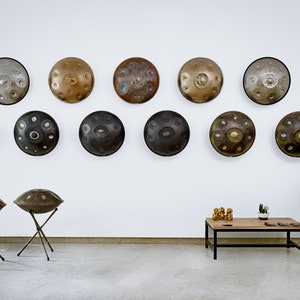 Puede incluir: Una colecci&oacute;n de 12 tambores de mano, tambi&eacute;n conocidos como tambores hang, se exhiben en una pared blanca. Dos handpans est&aacute;n en soportes en primer plano. Los handpans est&aacute;n hechos de metal y tienen una variedad de colores y acabados.