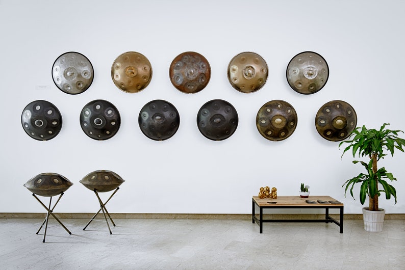 Puede incluir: Una colecci&oacute;n de 12 tambores de mano, tambi&eacute;n conocidos como tambores hang, se exhiben en una pared blanca. Dos handpans est&aacute;n en soportes en primer plano. Los handpans est&aacute;n hechos de metal y tienen una variedad de colores y acabados.