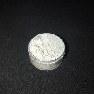 May include: A small, round container filled with a white, shimmery powder. The powder is likely used for crafting or cosmetic purposes.