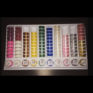 May include: A set of 14 small jars filled with different colored glitter. The jars are arranged in a white plastic tray. The jars are labeled with the color of the glitter inside. The labels read "Luster Pink", "Luster Pearls", "Luster Copper", "Luster Yellow", "Luster Green", "Luster Blue", "Luster Snow", "Luster Silver", "Luster Gold".