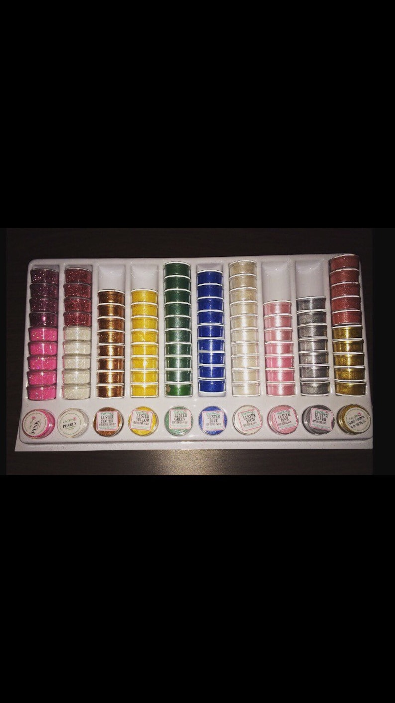 May include: A set of 14 small jars filled with different colored glitter. The jars are arranged in a white plastic tray. The jars are labeled with the color of the glitter inside. The labels read "Luster Pink", "Luster Pearls", "Luster Copper", "Luster Yellow", "Luster Green", "Luster Blue", "Luster Snow", "Luster Silver", "Luster Gold".