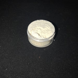 May include: A small, clear plastic container filled with a white powder. The container is sitting on a black surface.