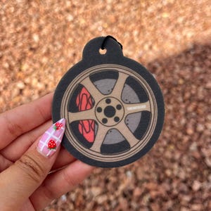 May include: Black car air freshener with a wheel design. The wheel is tan with a gray center and red brake calipers. The air freshener has a small hole at the top for hanging.