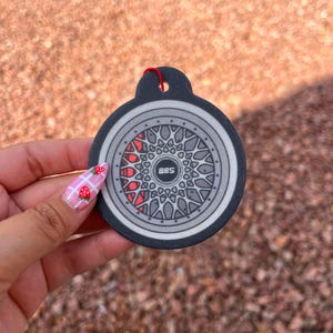 Puede incluir: Un dise&ntilde;o de rueda de coche redondo, negro y gris, con un acento rojo, con el texto "BBS" en el centro. El dise&ntilde;o est&aacute; en una etiqueta negra con una cuerda roja para colgar. El fondo es una superficie borrosa, de color marr&oacute;n rojizo.