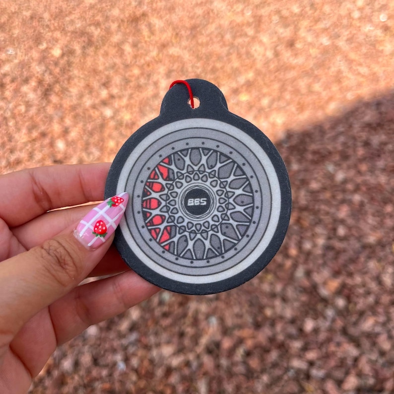 Puede incluir: Un dise&ntilde;o de rueda de coche redondo, negro y gris, con un acento rojo, con el texto "BBS" en el centro. El dise&ntilde;o est&aacute; en una etiqueta negra con una cuerda roja para colgar. El fondo es una superficie borrosa, de color marr&oacute;n rojizo.