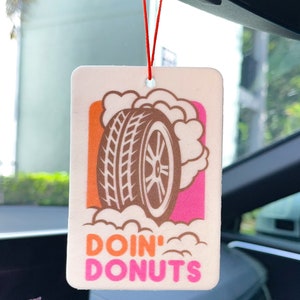 May include: Rectangular car air freshener with a tire graphic and the words "DOIN' DONUTS" in pink and orange. The air freshener is hanging from a red string in a car.