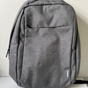 May include: A grey backpack with a textured fabric finish, featuring a large main compartment with a zipper closure, a smaller front zippered pocket, and a small rectangular Lenovo brand tag stitched onto the lower right corner of the front.