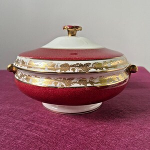Puede incluir: Un cuenco de servir cubierto con un exterior rojo moteado y detalles de hojas doradas. El cuenco tiene un borde y una tapa blancos, con un pomo dorado en la parte superior. El cuenco tiene dos asas doradas.