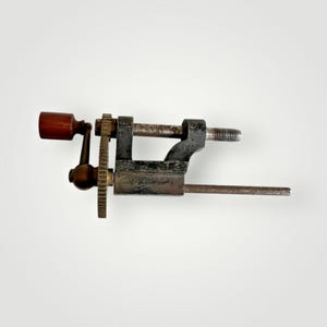 Puede incluir: Una herramienta de metal vintage con un mango de madera y un sistema de engranajes. La herramienta tiene una varilla de metal larga que se extiende desde el lateral.