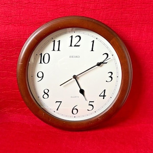 May include: A round wall clock with a dark brown wooden frame and a white face. The clock features black Arabic numerals from 1 to 12, black hour and minute hands, a thin black second hand, and the text Seiko and Quartz printed in black.