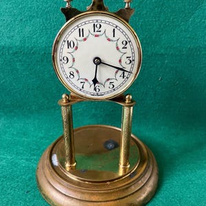 May include: A vintage brass clock with a round face featuring black numerals and floral accents. The clock has a decorative top and is supported by two ornate pillars on a circular base. The hands are black.