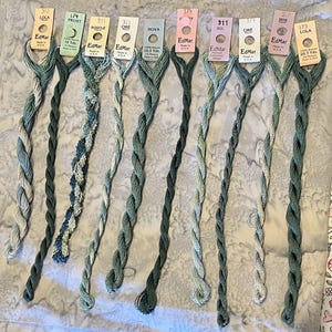 May include: Ten skeins of EdMar rayon embroidery thread in various shades of green, blue, and cream. Each skein is attached to a labeled paper tag displaying the brand name, color number, and length in yards, ranging from 10.5 to 20.7 yards.