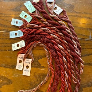 May include: A collection of colorful embroidery floss in shades of red, brown, and tan, with attached labels. The labels display the brand name "EdMar" and various color names and numbers, including "Frost", "Nova", and "Glory".