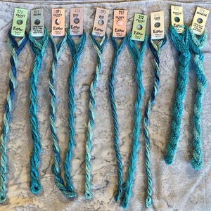 May include: Ten skeins of EdMar rayon embroidery thread in various shades of blue and teal. Each skein is attached to a paper tag labeled with the brand name, color number, and style name such as Frost, Nova, Glory, Iris, Cire, or Boucle.