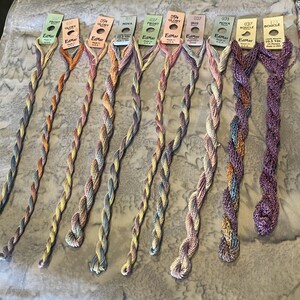 May include: Ten skeins of EdMar embroidery thread in various variegated colorways, including shades of blue, yellow, pink, and purple. Each skein features a paper tag at the top with brand name, color number, and length in yards.