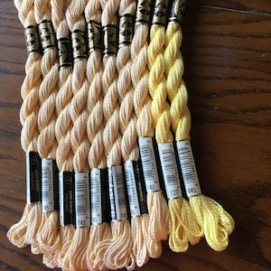 May include: A collection of 10 spools of DMC embroidery floss in shades of yellow. The spools are labeled with the DMC color numbers 745, 727, and 445. The spools are arranged in a row, with the yellowest shade at the top and the lightest shade at the bottom.