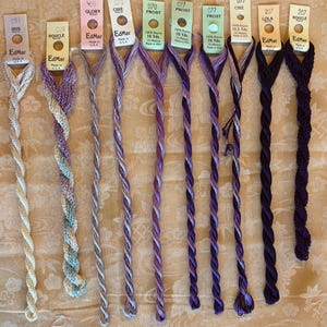 May include: A collection of nine EdMar rayon embroidery threads in various shades of purple, lavender, and white. Each thread is attached to a labeled paper tag displaying the brand name, color number, and product type like Iris, Boucle, or Glory.