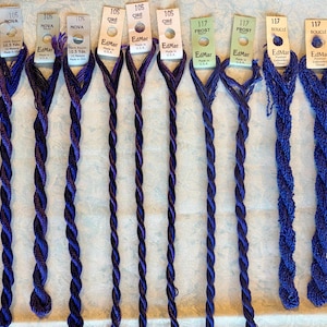 May include: Ten skeins of EdMar embroidery thread arranged in a row. The first three are labeled 105 Nova, the next three 105 Cire, the next two 117 Frost, and the final two 117 Boucle. Each skein features a purple or blue twisted rayon thread.