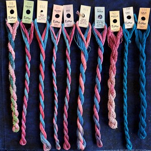 May include: Ten skeins of EdMar rayon embroidery thread arranged vertically. The threads feature various twisted textures and color combinations, including shades of pink, blue, and green. Each skein has a paper tag with brand and color information.
