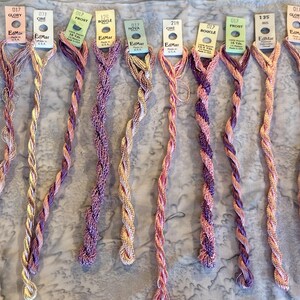 May include: Nine skeins of EdMar embroidery thread arranged in a row, each with a paper label at the top. The threads feature various twisted textures and color combinations, including shades of pink, purple, yellow, and metallic tones, each labeled with specific product codes like 017, 031, 125, 218, and 135.