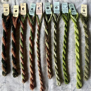 May include: Nine skeins of EdMar rayon embroidery thread arranged in a row, each with a paper tag displaying brand name, color number, and length in yards. The threads feature various twisted textures and shades of green, brown, and copper tones.