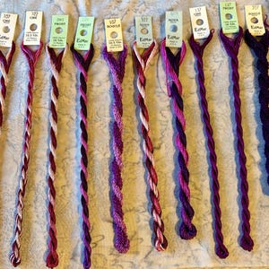 May include: Nine skeins of EdMar embroidery thread arranged in a row, featuring various shades of pink, magenta, and deep purple. Each skein is attached to a white paper tag displaying the brand name, color number, style name, and length in yards.