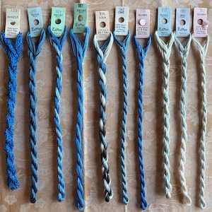 May include: Ten skeins of EdMar rayon embroidery thread arranged in a row, featuring various shades of blue and cream. Each skein is attached to a paper tag labeled with a color name, number, and length in yards or meters, made in the USA.