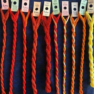May include: Ten skeins of EdMar rayon embroidery thread in shades of orange, red, and yellow, each with a paper label detailing the brand, color name, number, and length in yards or meters, arranged vertically in a row against a dark blue background.