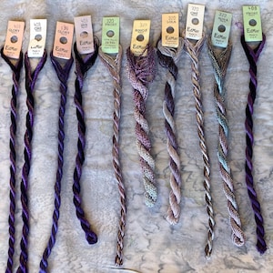 May include: Nine skeins of EdMar embroidery thread in various purple, lavender, and metallic shades. Each skein features a paper tag with brand name, color number, and style name like Glory, Cire, Iris, Frost, Boucle, Lola, and Nova, made in the USA.