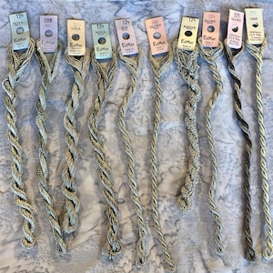 May include: Ten strands of EdMar embroidery thread in various metallic shades, each attached to a labeled paper tag. The threads feature twisted textures and include labels such as Nova, Iris, Lola, Glory, Bouclé, and Cire, with some tags noting 100% Rayon.
