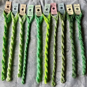May include: Nine skeins of EdMar embroidery thread in various shades of green, ranging from light lime to deep forest green. Each skein is attached to a paper tag labeled with brand name, color number, style name, and length in yards.