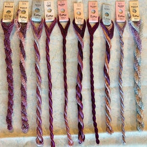 May include: Ten strands of EdMar embroidery thread in various colors and textures, each attached to a labeled paper tag. The tags display brand names like Bouclé, Nova, Ciré, Glory, and Lola, along with color numbers and 10.5 yards of rayon.