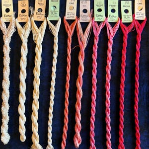 May include: Ten skeins of EdMar rayon embroidery thread arranged in a row, featuring a gradient from cream and beige to coral, pink, and red. Each skein has a paper tag with the brand name, color number, name, and length in yards or meters.