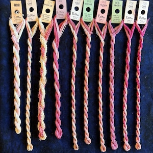 May include: Ten skeins of EdMar rayon embroidery thread in various shades of pink, cream, and coral. Each skein features a paper tag at the top with brand name, color name, and length in yards. The threads are twisted and arranged vertically.