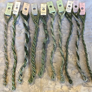 May include: Nine skeins of green embroidery thread arranged vertically, each with a paper label at the top. Labels include brand name EdMar and color names like Frost, Lola, Nova, Boucle, Iris, and Glory, with lengths listed in yards and meters.