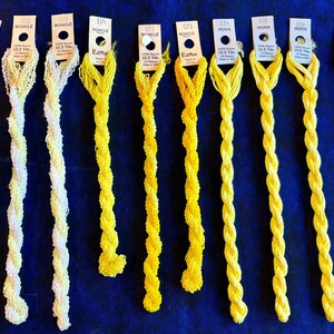 May include: Ten skeins of rayon embroidery thread arranged in a row, ranging from pale cream to vibrant yellow. Each skein features a paper tag labeled with brand names Nova or Bouclé, color numbers, and the text 10.5 Yds. Made in USA.