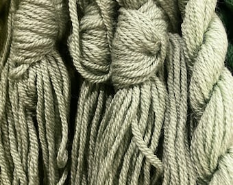 May include: Close-up of multiple skeins of soft, sage green yarn. The yarn is arranged in a loose, tangled pile, with some strands tied together in knots.