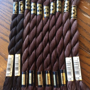 May include: A collection of ten spools of DMC embroidery floss in shades of brown. The spools are labeled with the DMC numbers 3371, 838, 938, and 738.