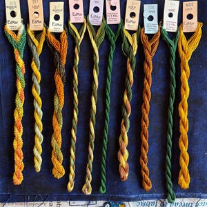 May include: Ten skeins of EdMar embroidery thread in various shades of green, yellow, and orange, each attached to a labeled paper tag. The threads are twisted and arranged vertically, displaying textures ranging from smooth to bouclé.