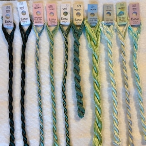 May include: A collection of ten EdMar rayon embroidery threads in various green and black shades, each with a paper label displaying the brand name, color number, and length in yards or meters, arranged vertically on a white surface.