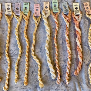 May include: Ten skeins of EdMar rayon embroidery thread in various shades of peach, cream, and light orange. Each skein features a paper tag with the brand name, color name, and length in yards, such as 10.5 Yds or 16 Yds, displayed in a vertical row.