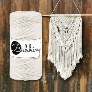 Op de afbeelding: Een rol natuurlijke katoenen koord, met het label "Bobbiny", naast een witte macrame wandhanger met een geometrisch patroon.