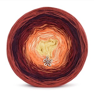 Op de afbeelding: Een bol ombre garen in tinten rood, oranje en geel. Het garen is opgerold in een bolvorm en heeft een zilveren hanger eraan.