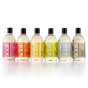 May include: Six clear glass bottles with white labels and black caps. The bottles are filled with a clear liquid and have different colored designs on the labels. The labels read "soak" and have different scents listed: "cucumber", "yuzu", "pineapple", "fig", "white tea", and "honey".