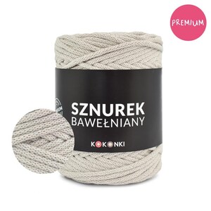 Może przedstawiać: Cylindryczna szpula beżowego sznurka bawełnianego z czarną etykietą. Etykieta zawiera napis "SZUREK BAWEŁNIANY" oraz nazwę marki "KOKONKI". Różowe koło z napisem "PREMIUM" znajduje się w prawym górnym rogu. Z lewej strony u dołu zbliżenie sznurka.