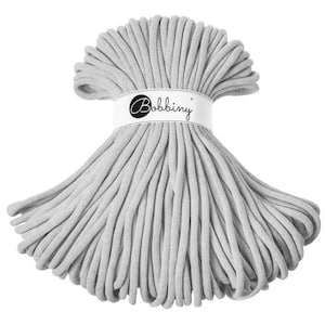 Può includere: Un grande rotolo di corda di cotone grigio chiaro, perfetto per progetti di artigianato. La corda è impilata ordinatamente e legata con un'etichetta bianca che recita "Bobbiny".