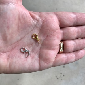 May include: Two metal lobster claw clasps, one gold-colored and one silver-colored, are displayed. The clasps are small, with a spring-loaded mechanism for secure closure. They are likely used for jewelry making or crafting.