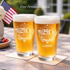 May include: Two pint glasses filled with beer, featuring a patriotic design with the text "Fifty States, One Nation" and "We The People 1776-2026". One glass is personalized with the name "GUNNER". The image includes an American flag.