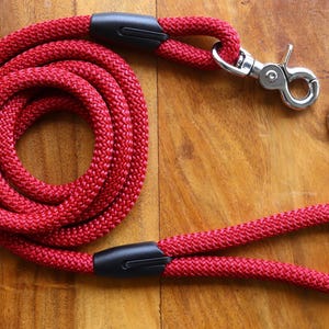 May include: Red rope dog leash with a black handle and a silver clip. The leash is coiled up on a wooden surface.